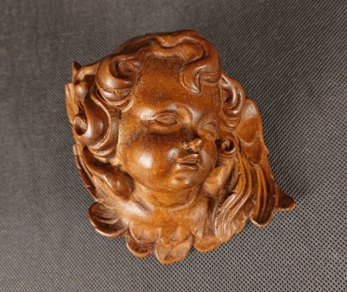 Rzeźba przedstawia ładną drewnianą główkę amorka z drobno wyrzeźbionymi lokami, patrzący w lewo. Pod brodą zaznaczono kokardę, a po bokach delikatne skrzydełka. Gładka, ciepła faktura drewna podkreśla ręczne dłuto.