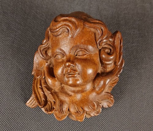 Rzeźba przedstawia drewnianą główkę amorka z drobno wyrzeźbionymi lokami i zamkniętymi oczami. Pod brodą zaznaczono kokardę, a po bokach delikatne skrzydełka. Gładka, ciepła faktura drewna podkreśla ręczne dłuto.