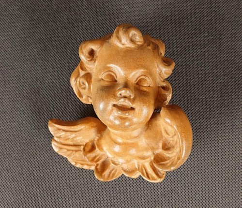 Jasna, drewniana rzeźba przedstawia uskrzydloną główkę. Ma okrągłą twarz z zamkniętymi oczami i lekko uśmiechniętymi ustami. Widoczne są kręcone włosy i dwa małe skrzydła z zaznaczonymi piórami.