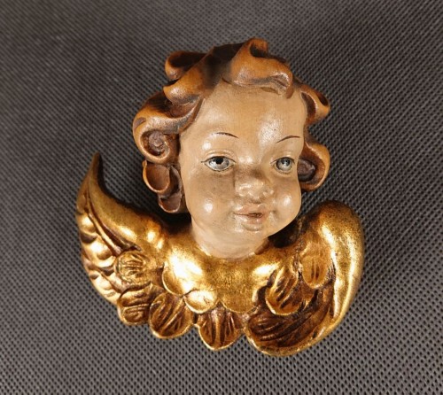 Ręcznie wyrzeźbiona drewniana, polichromowana główka amorka ze skrzydełkami, z bujnymi lokami i złoconymi skrzydełkami przy policzkach. Twarz o lekko rozchylonych ustach i figlarnym spojrzeniu.