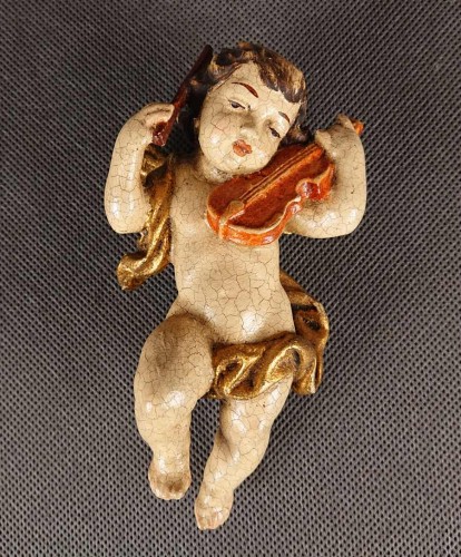 Prezentowana rzeźba to misternie wykonany, polichromowany cherubin – klasyczny putto z ikonografii barokowej – unoszący się w powietrzu podczas wykonywania muzycznego koncertu. Całość wyrzeźbiona jest z drewna o delikatnej, lekko pękającej powierzchni.