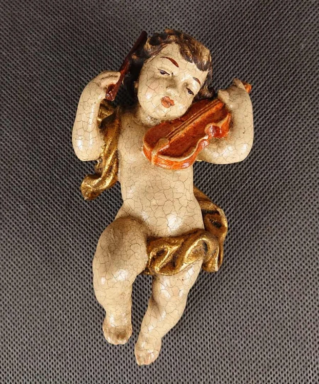 Prezentowana rzeźba to misternie wykonany, polichromowany cherubin – klasyczny putto z ikonografii barokowej – unoszący się w powietrzu podczas wykonywania muzycznego koncertu. Całość wyrzeźbiona jest z drewna o delikatnej, lekko pękającej powierzchni.
