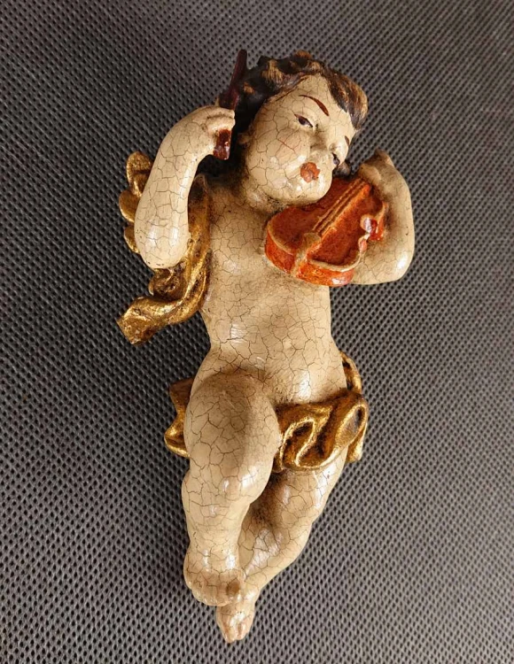 Prezentowana rzeźba to misternie wykonany, polichromowany cherubin – klasyczny putto z ikonografii barokowej – unoszący się w powietrzu podczas wykonywania muzycznego koncertu. Całość wyrzeźbiona jest z drewna o delikatnej, lekko pękającej powierzchni.