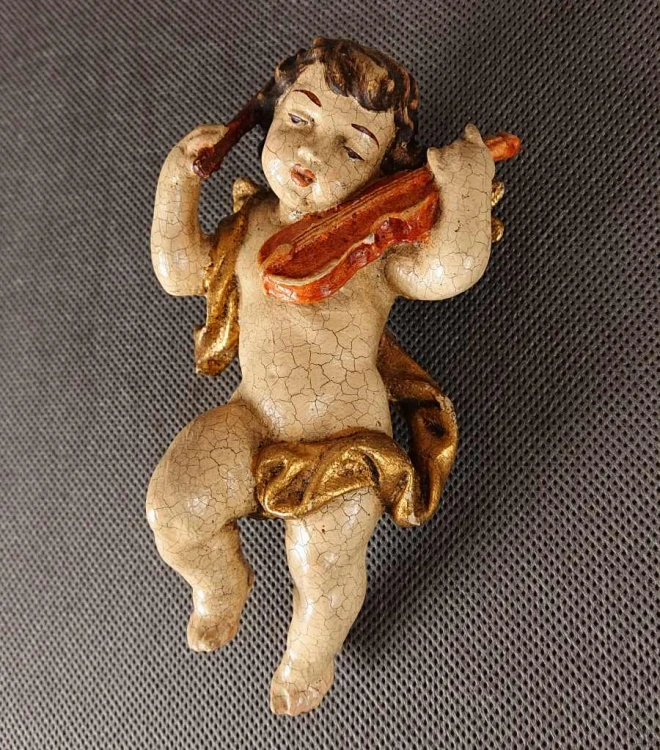 Prezentowana rzeźba to misternie wykonany, polichromowany cherubin – klasyczny putto z ikonografii barokowej – unoszący się w powietrzu podczas wykonywania muzycznego koncertu. Całość wyrzeźbiona jest z drewna o delikatnej, lekko pękającej powierzchni.