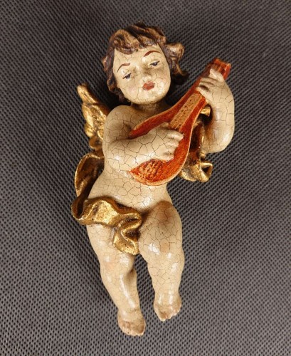 Przedstawiona rzeźba to niewielki, lecz niezwykle ekspresyjny putto – amorkowy posłaniec mitologii – zawieszony w ruchu podczas gry na lutni. Wykonany z drewna rzeźbiarski korpus emanuje autentycznym patynowaniem starej polichromii