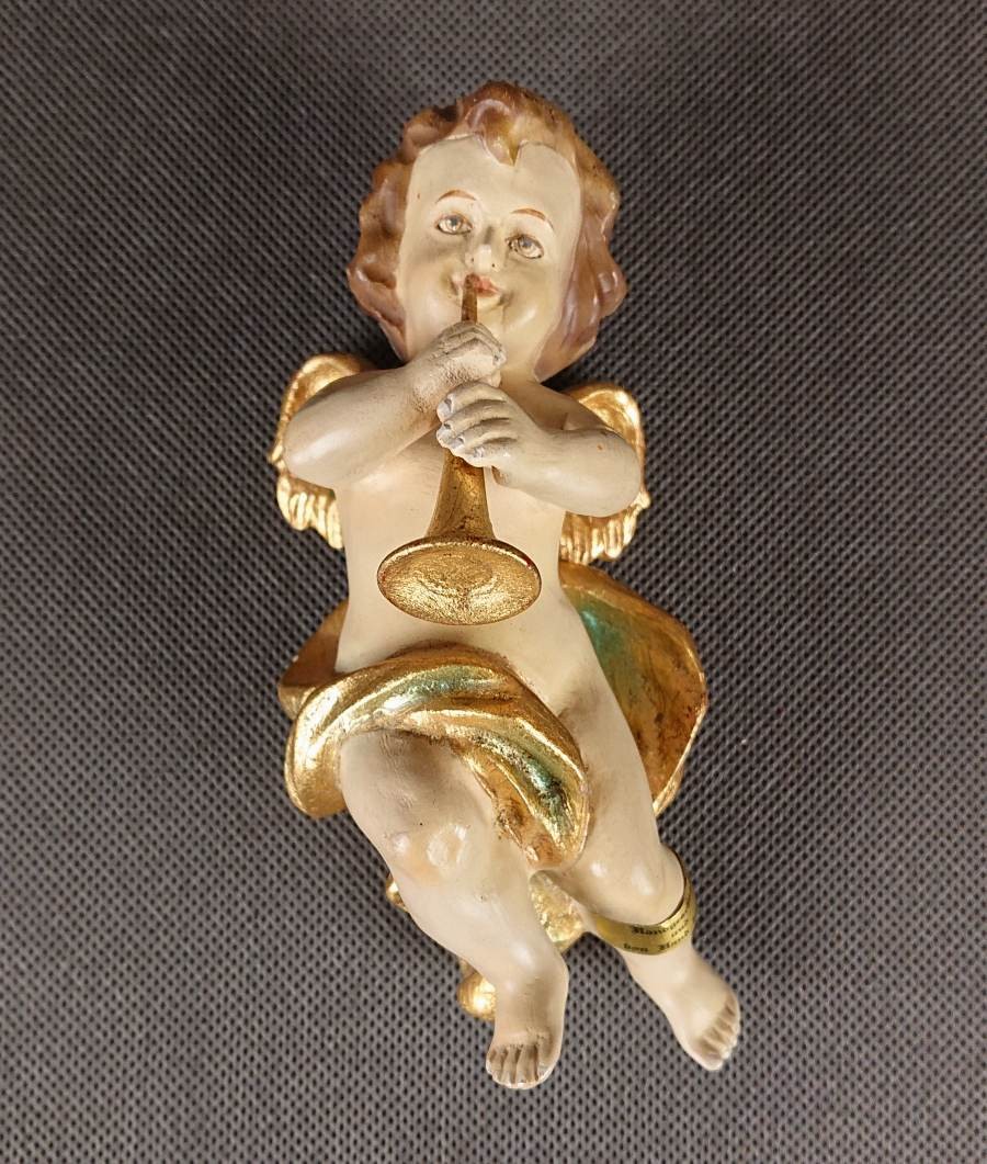 Polichromowana rzeźba z drewna przedstawia amorka ze skrzydełkami, unoszącego się i grającego na złotej trąbce. Jego ciało ma jasną, cielistą barwę. Włosy, w odcieniach brązu i złota, opadają mu lokami na ramiona