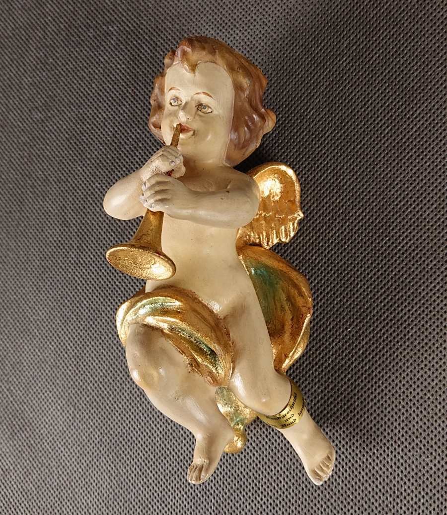 Polichromowana rzeźba z drewna przedstawia amorka ze skrzydełkami, unoszącego się i grającego na złotej trąbce. Jego ciało ma jasną, cielistą barwę. Włosy, w odcieniach brązu i złota, opadają mu lokami na ramiona