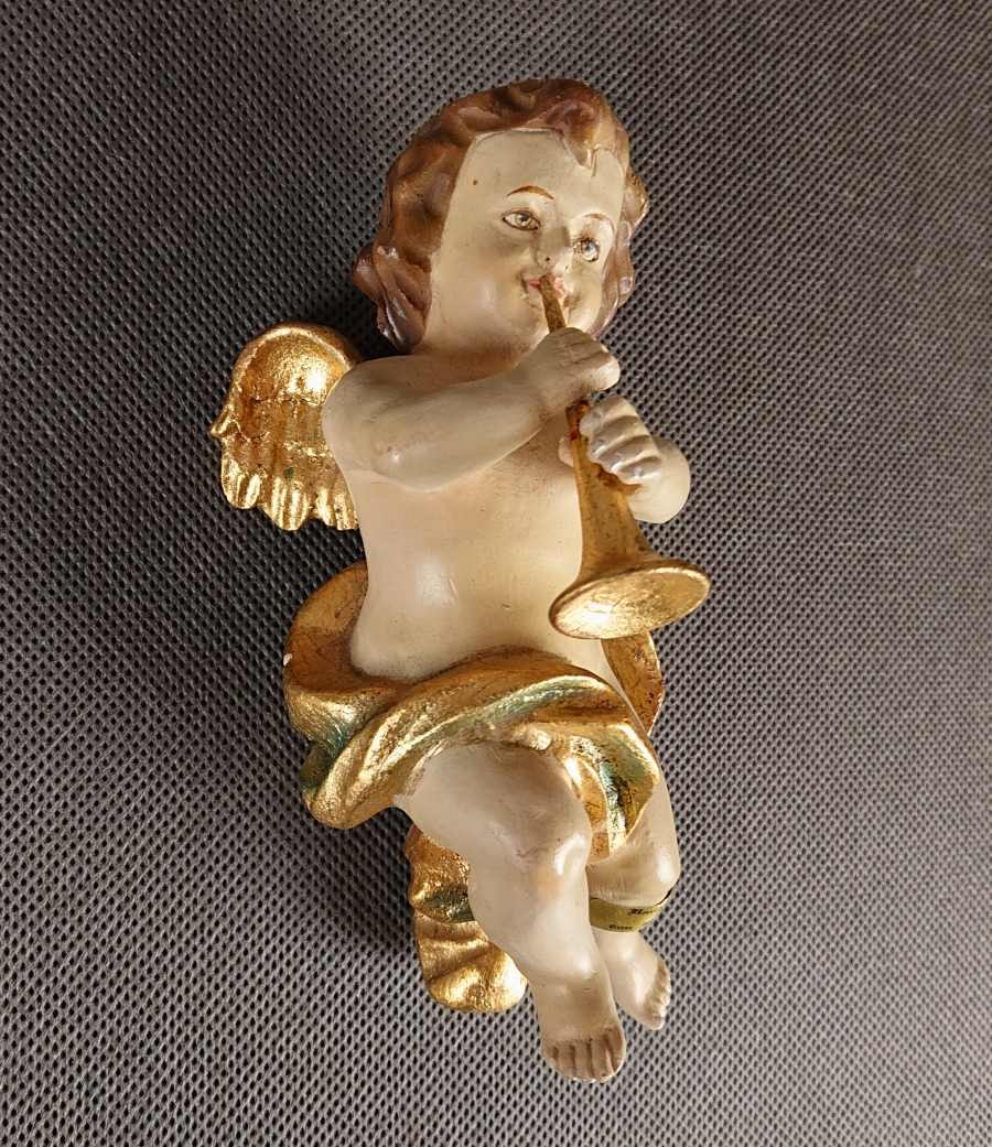 Polichromowana rzeźba z drewna przedstawia amorka ze skrzydełkami, unoszącego się i grającego na złotej trąbce. Jego ciało ma jasną, cielistą barwę. Włosy, w odcieniach brązu i złota, opadają mu lokami na ramiona
