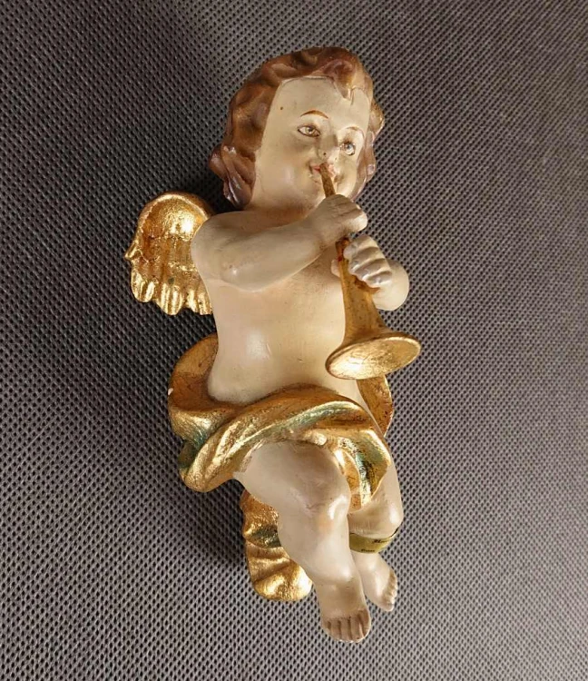 Polichromowana rzeźba z drewna przedstawia amorka ze skrzydełkami, unoszącego się i grającego na złotej trąbce. Jego ciało ma jasną, cielistą barwę. Włosy, w odcieniach brązu i złota, opadają mu lokami na ramiona