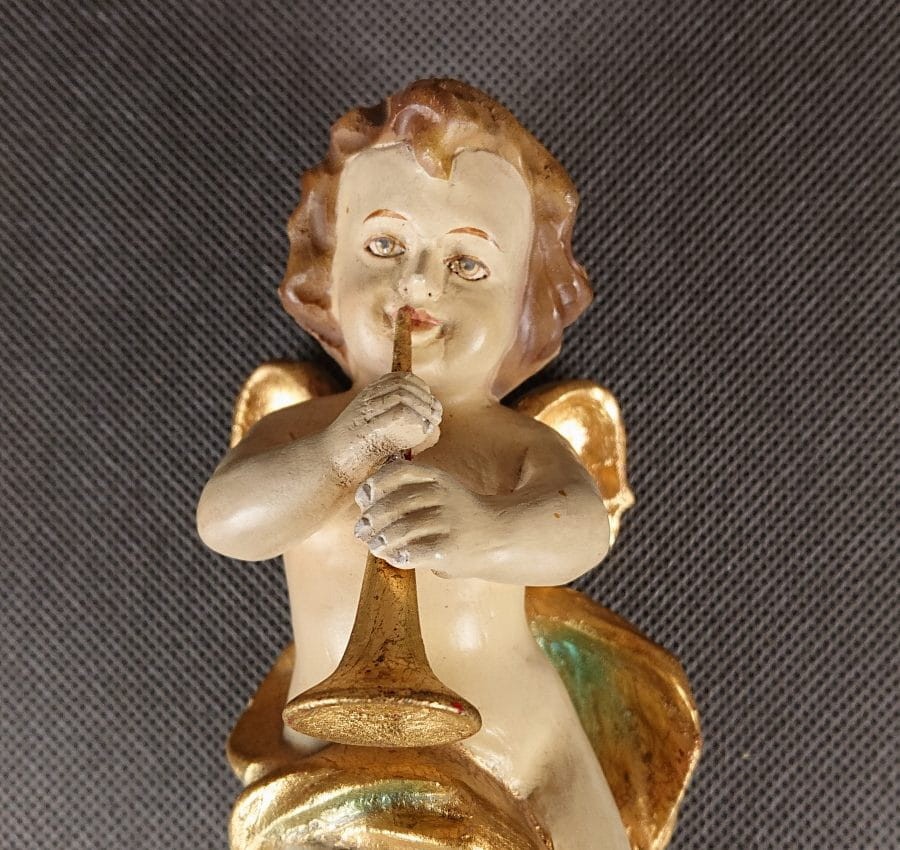 Polichromowana rzeźba z drewna przedstawia amorka ze skrzydełkami, unoszącego się i grającego na złotej trąbce. Jego ciało ma jasną, cielistą barwę. Włosy, w odcieniach brązu i złota, opadają mu lokami na ramiona