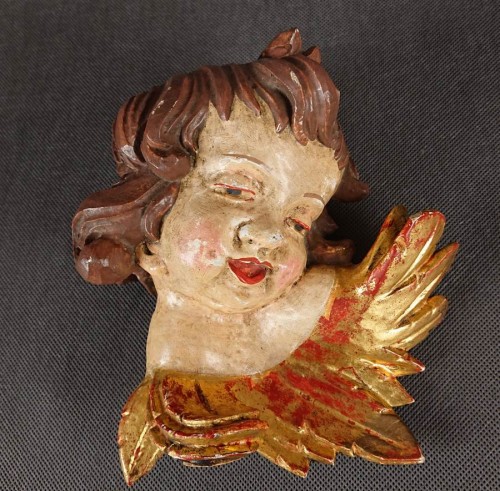 Duża polichromowana głowa amora wyrzeźbiona z drewna w XIX w. Postać przedstawiono z pulchną, dziecięcą twarzą o delikatnie różowawych policzkach i pełnych ustach, zwróconą w lewo. Gęste loki podkreślono ciemnobrązową polichromią, skrzydełka złoconą farbą
