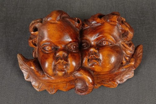 Rzeźba wykonana z ciepłego, polerowanego drewna przedstawia dwie złączone główki amorów. Pulchne, dziecięce twarze o delikatnie wyrazistych rysach – zaokrąglonych policzkach, subtelnie zarysowanych ustach i dużych, pełnych ciekawości oczach