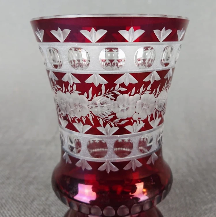 Ta szklanka w stylu Biedermeier, sygnowana "E. Krause 91", prezentuje charakterystyczną dla tego okresu elegancję i kunszt wykonania. Wykonana jest z rubinowego szkła, które swoją głęboką, czerwoną barwą przyciąga wzrok.