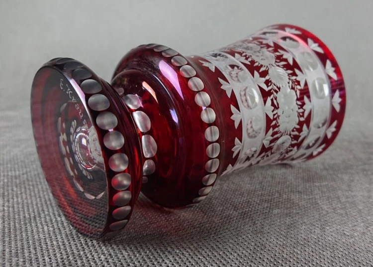 Ta szklanka w stylu Biedermeier, sygnowana "E. Krause 91", prezentuje charakterystyczną dla tego okresu elegancję i kunszt wykonania. Wykonana jest z rubinowego szkła, które swoją głęboką, czerwoną barwą przyciąga wzrok.