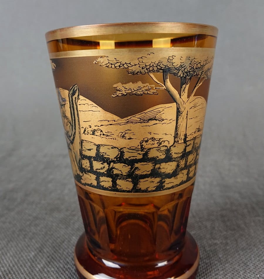 Szklanka, wykonana i ręcznie dekorowana w latach 20-30 XX wieku, prezentuje bogate zdobienie charakterystyczne dla tego okresu. Wykonana jest z miodowego, szkła, które w górnej partii jest matowione, tworząc kontrastującą powierzchnię dla dekoracji.