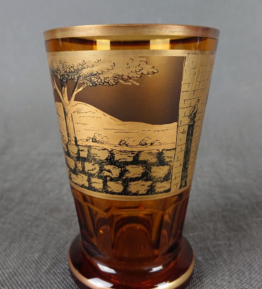 Szklanka, wykonana i ręcznie dekorowana w latach 20-30 XX wieku, prezentuje bogate zdobienie charakterystyczne dla tego okresu. Wykonana jest z miodowego, szkła, które w górnej partii jest matowione, tworząc kontrastującą powierzchnię dla dekoracji.