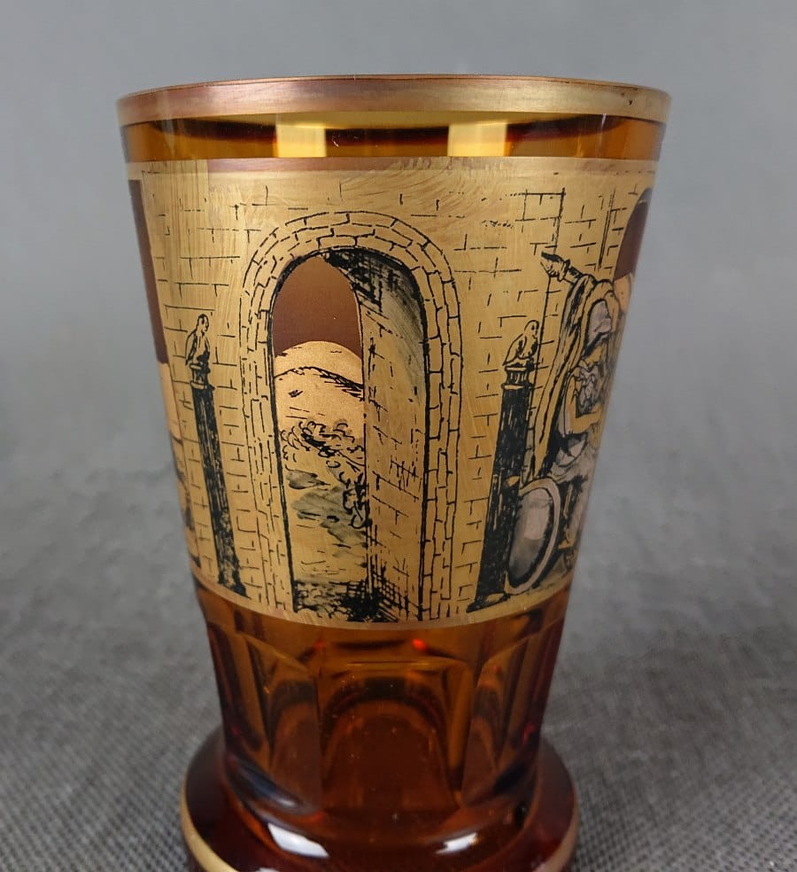 Szklanka, wykonana i ręcznie dekorowana w latach 20-30 XX wieku, prezentuje bogate zdobienie charakterystyczne dla tego okresu. Wykonana jest z miodowego, szkła, które w górnej partii jest matowione, tworząc kontrastującą powierzchnię dla dekoracji.