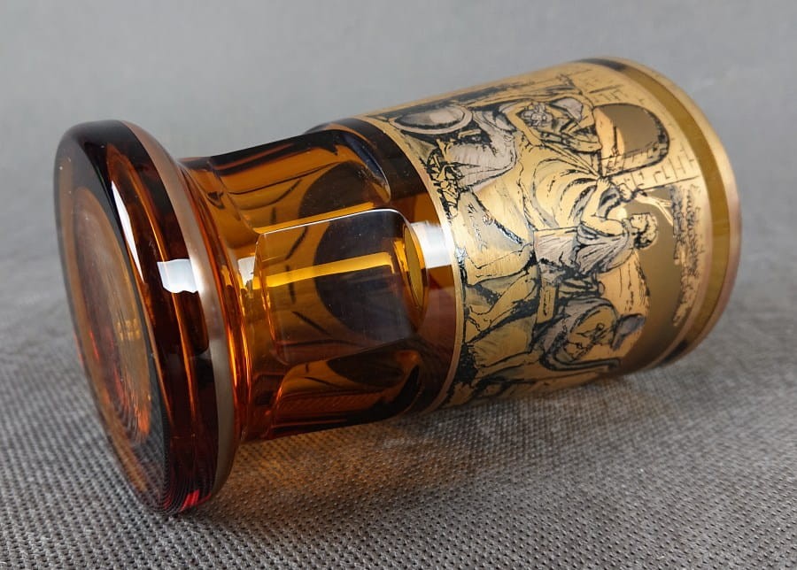 Szklanka, wykonana i ręcznie dekorowana w latach 20-30 XX wieku, prezentuje bogate zdobienie charakterystyczne dla tego okresu. Wykonana jest z miodowego, szkła, które w górnej partii jest matowione, tworząc kontrastującą powierzchnię dla dekoracji.