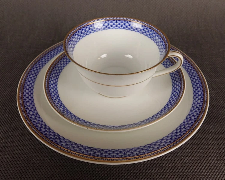 Zestaw śniadaniowy z fabryki porcelany C.M.Hutschenreuther składa się z filiżanki, spodka i talerza deserowego. Wszystkie elementy wykonane są z białej, szkliwionej porcelany i charakteryzują się geometrycznym zdobieniem w odcieniach błękitu i złota.