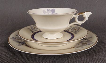 Przedwojenny zestaw śniadaniowy z niemieckiej fabryki porcelany Hertel Jacob. składa się z filiżanki, spodka i talerza deserowego. Zestaw charakteryzuje się elegancką, kremową porcelaną oraz delikatnymi zdobieniami w odcieniach błękitu, szarości i złota.