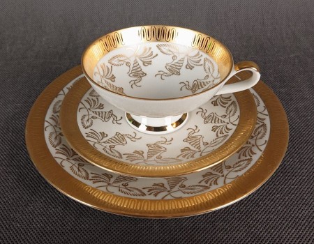 Porcelanowy zestaw śniadaniowy z niemieckiej fabryki Hertel Jacob & Co. to elegancki komplet składający się z filiżanki, spodka i talerza deserowego. Wykonana z białej, szkliwionej porcelany i bogato zdobiona złotymi, reliefowymi i malowanymi wzorami.
