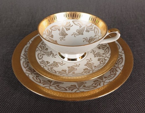 Porcelanowy zestaw śniadaniowy z niemieckiej fabryki Hertel Jacob & Co. to elegancki komplet składający się z filiżanki, spodka i talerza deserowego. Wykonana z białej, szkliwionej porcelany i bogato zdobiona złotymi, reliefowymi i malowanymi wzorami.