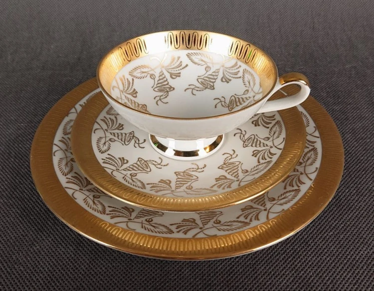 Porcelanowy zestaw śniadaniowy z niemieckiej fabryki Hertel Jacob & Co. to elegancki komplet składający się z filiżanki, spodka i talerza deserowego. Wykonana z białej, szkliwionej porcelany i bogato zdobiona złotymi, reliefowymi i malowanymi wzorami.
