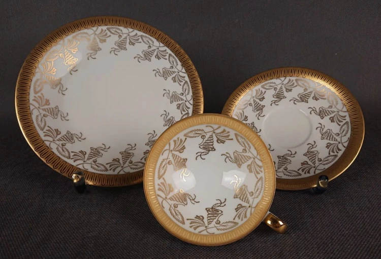 Porcelanowy zestaw śniadaniowy z niemieckiej fabryki Hertel Jacob & Co. to elegancki komplet składający się z filiżanki, spodka i talerza deserowego. Wykonana z białej, szkliwionej porcelany i bogato zdobiona złotymi, reliefowymi i malowanymi wzorami.