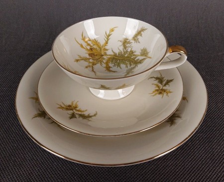Zestaw śniadaniowy z niemieckiej fabryki porcelany Hertel Jacob & Co. to elegancki komplet składający się z filiżanki, spodka i talerza deserowego. Elementy wykonane są z białej, szkliwionej porcelany i zdobione motywami ostów i złotymi akcentami.