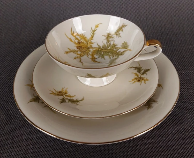 Zestaw śniadaniowy z niemieckiej fabryki porcelany Hertel Jacob & Co. to elegancki komplet składający się z filiżanki, spodka i talerza deserowego. Elementy wykonane są z białej, szkliwionej porcelany i zdobione motywami ostów i złotymi akcentami.