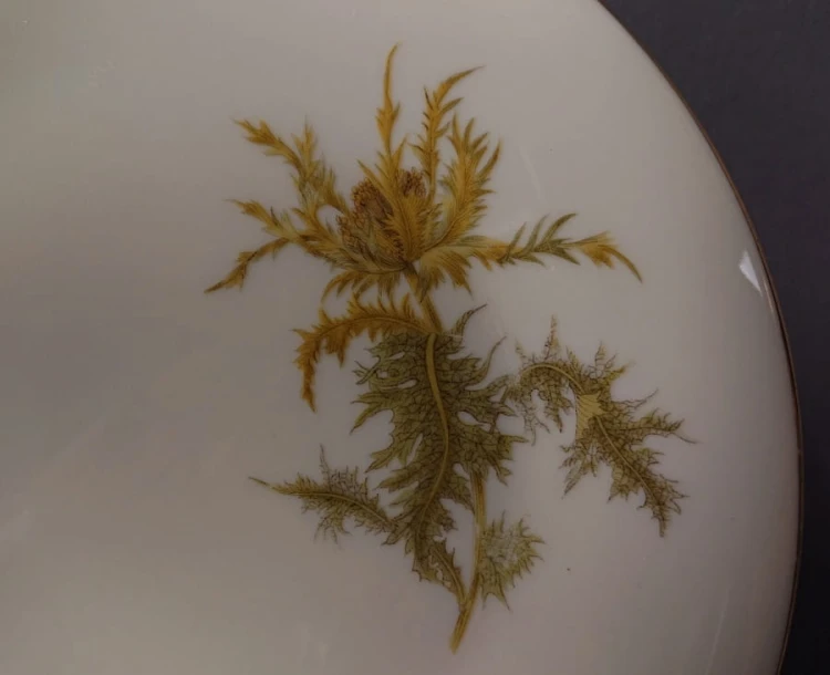 Zestaw śniadaniowy z niemieckiej fabryki porcelany Hertel Jacob & Co. to elegancki komplet składający się z filiżanki, spodka i talerza deserowego. Elementy wykonane są z białej, szkliwionej porcelany i zdobione motywami ostów i złotymi akcentami.