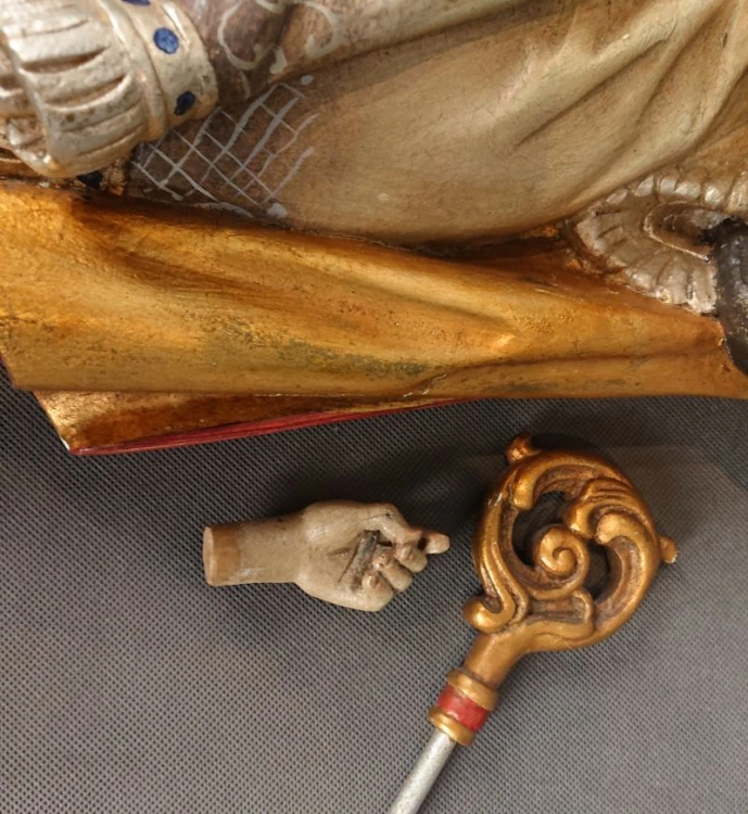 Zbliżenie na dół szaty i odłączoną drewnianą lewą dłoń leżącą obok złoconej głowicy pastorału.