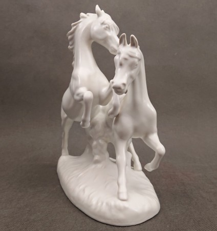 Porcelanowa figura białego, lśniącego konia marki Goebel na jasnym tle, ujęcie zprzodu eksponujące sylwetkę zwierzęcia.
