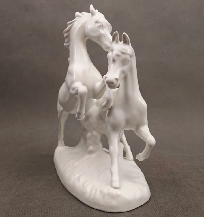 Porcelanowa figura białego, lśniącego konia marki Goebel na jasnym tle, ujęcie zprzodu eksponujące sylwetkę zwierzęcia.