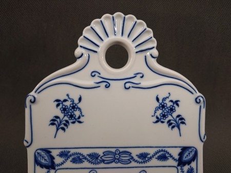 Widok na górną część porcelanowej deski z faliście wyciętym, ozdobnym uchwytem i okrągłym otworem do zawieszenia, zdobioną dwoma symetrycznymi bukietami kwiatów.