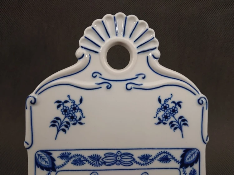 Widok na górną część porcelanowej deski z faliście wyciętym, ozdobnym uchwytem i okrągłym otworem do zawieszenia, zdobioną dwoma symetrycznymi bukietami kwiatów.