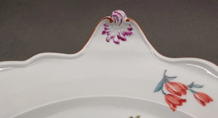 Zbliżenie na wewnętrzną krawędź owalnego półmiska z porcelany, widoczne drobne fioletowe i niebieskie kwiatki oraz terakotowa linia na faliście wyciętym rancie.