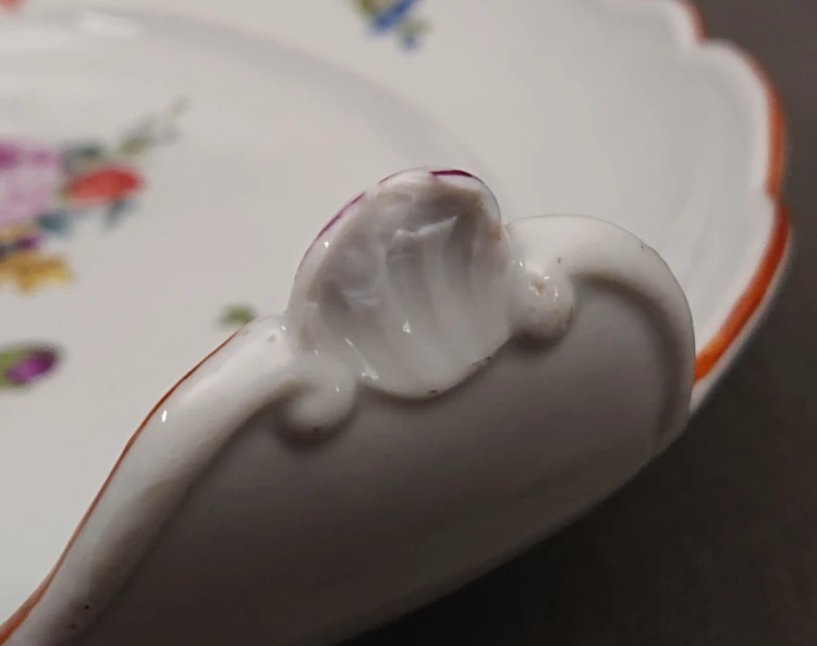Detal spodu uniesionego uszka porcelanowego, ukazujący powierzchniowy, dawny ubytek masy porcelanowej na dolnej krawędzi reliefu.