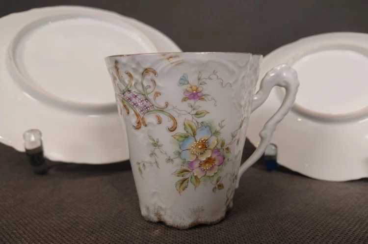 Zbliżenie na profil filiżanki z kwiatowym malunkiem i złoceniami na tle białych naczyń porcelanowych z końca XIX wieku.